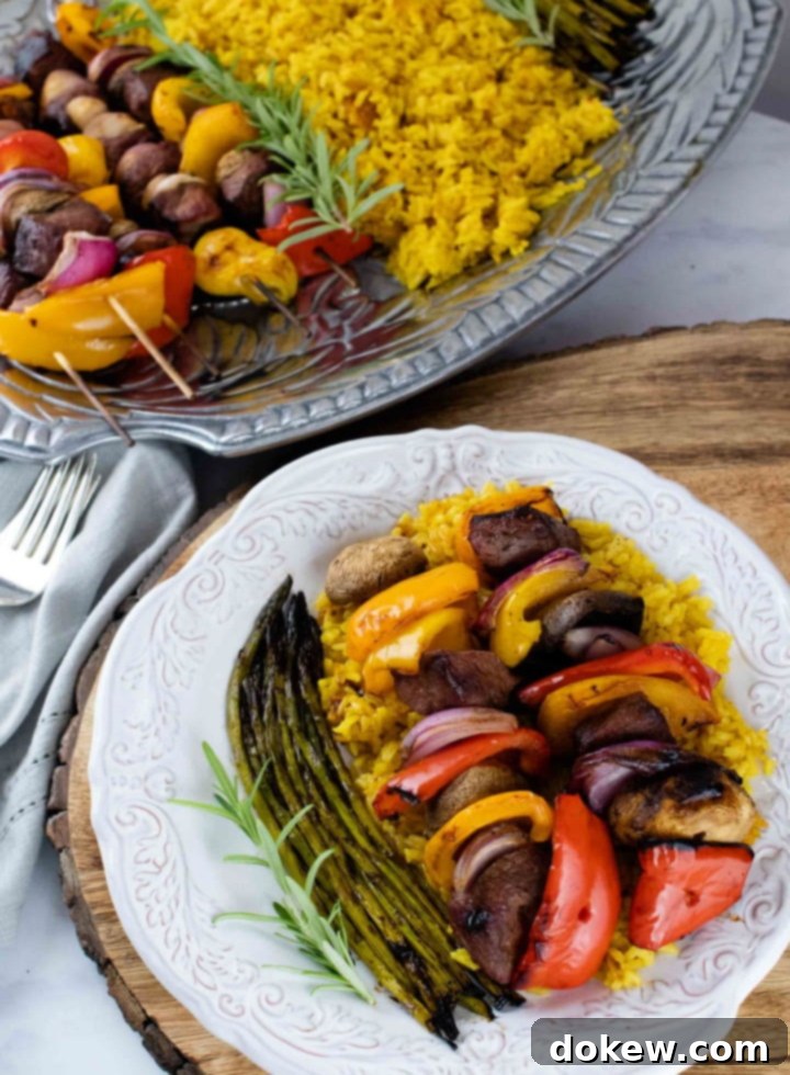 beef-vegetable-kabobs-post-vert-min grilled kabobs of red, yellow, and orange bell pepper pieces alternated with mushrooms, red onion pieces, and marinated beef and layered over yellow rice with grilled asparagus on a white plate with a silver fork with a serving platter of kabobs, yellow rice, and asparagus to the side