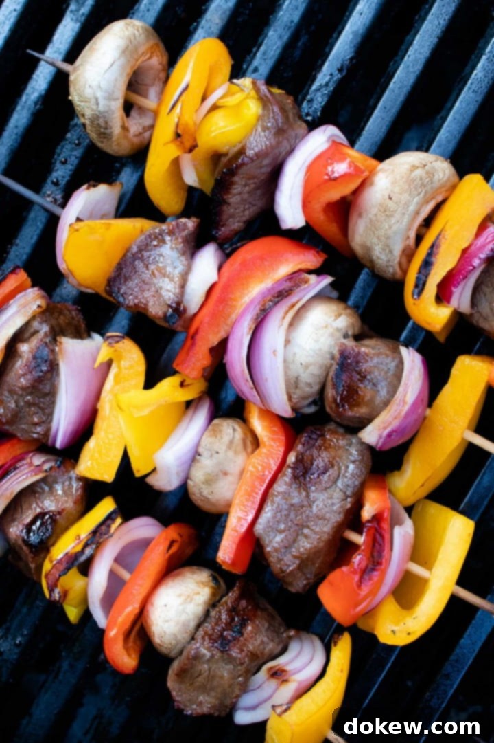 beef-vegetable-kabobs-ip-1-min kabobs of red, yellow, and orange bell pepper pieces alternated with mushrooms, red onion pieces, and marinated beef cooking on a grill with visible char marks