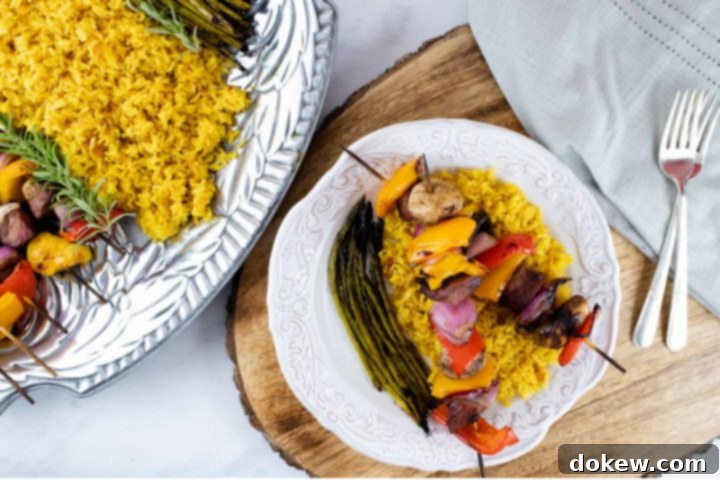 beef-vegetable-kabobs-ip-5-min grilled kabobs of red, yellow, and orange bell pepper pieces alternated with mushrooms, red onion pieces, and marinated beef and layered over yellow rice with grilled asparagus on a white plate with a silver fork with a serving platter of kabobs, yellow rice, and asparagus to the side