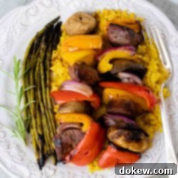 Tender Beef Skewers and Aromatic Yellow Rice 2 grilled kabobs of red, yellow, and orange bell pepper pieces alternated with mushrooms, red onion pieces, and marinated beef and layered over yellow rice with grilled asparagus on a white plate with a silver fork