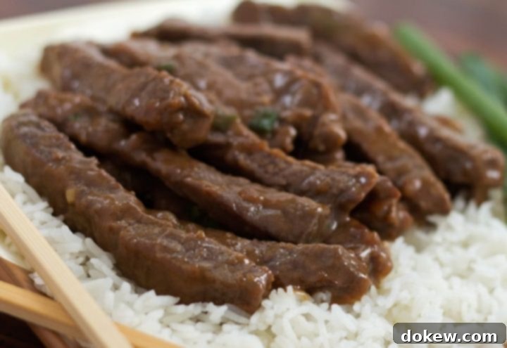 Mongolian Beef Stir Fry Sliced steak with a sauce on a bed of rice with scallions in the background and chopsticks in the foreground.
