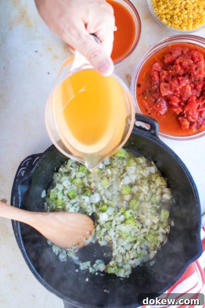 Pasta e Fagioli A measuring cup of broth is being poured into a cast iron stew pot of chopped celery and onions. A wooden spoon is stirring the contents of the pan. At the top right of the photo is a clear glass bowl with roasted tomatoes in tomato juice.