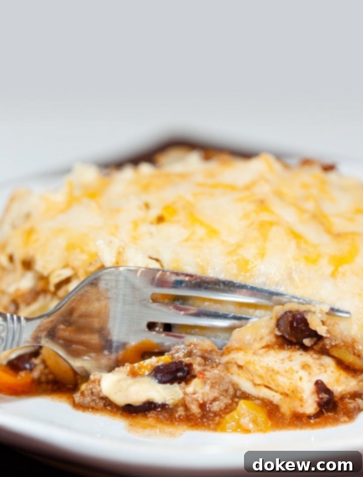 Beefy Bean Bake A tortilla covered with melted cheese, on top of ground beef, black beans, and tomatoes, all on a white plate. A fork is cutting into the tortilla.