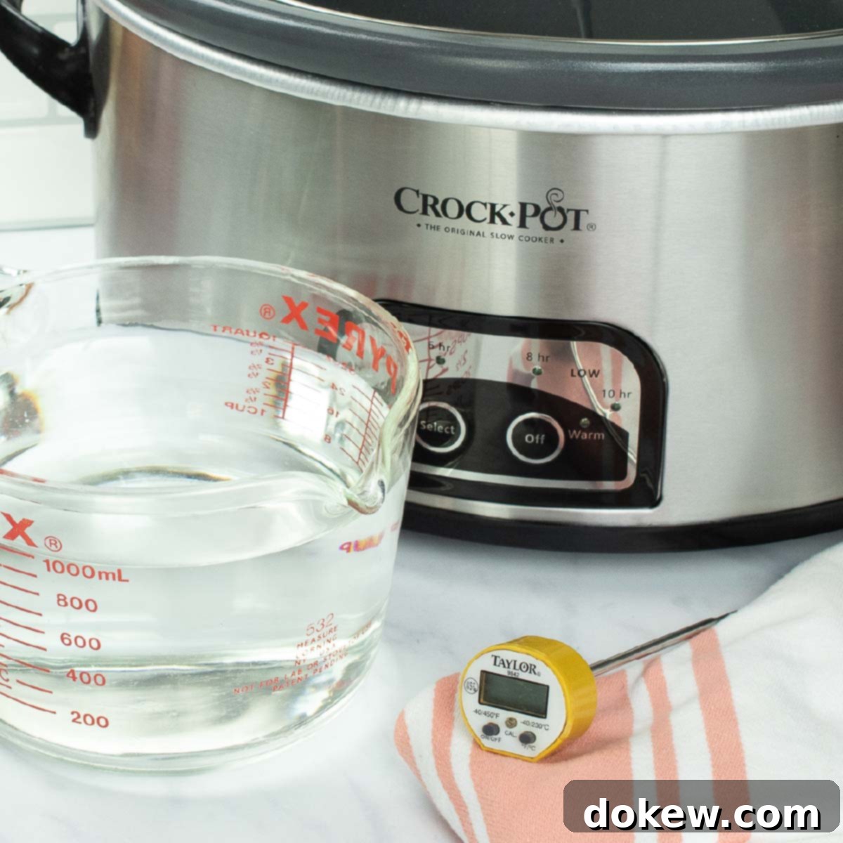 A modern slow cooker with a glass lid, a measuring cup of water, and a food thermometer positioned nearby, ready for a performance test.