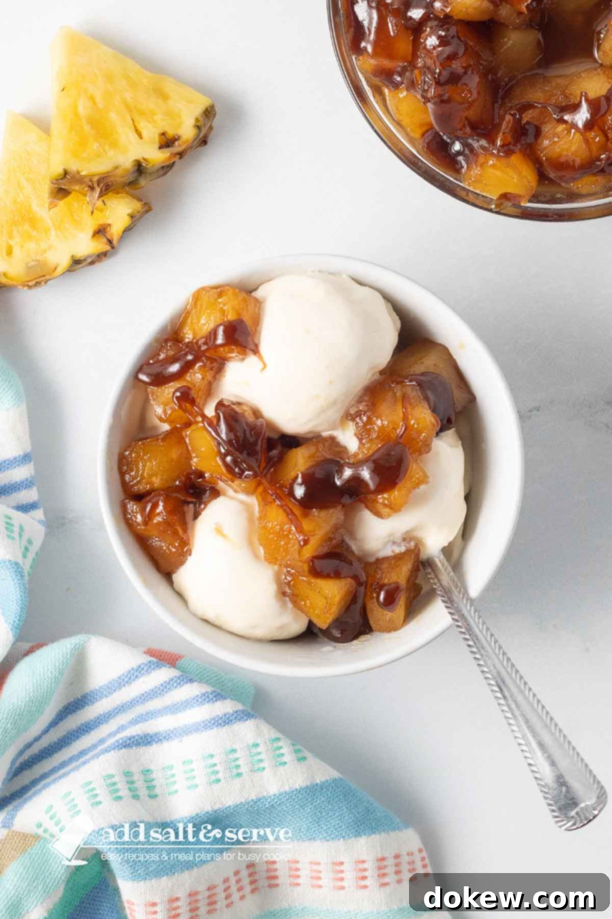 Sweet Golden Caramelized Pineapple Just 3 Ingredients 5 Bowl of caramelized pineapple chunks and vanilla ice cream drizzled with caramel sauce beside a tea towel and sliced pineapple on a counter.