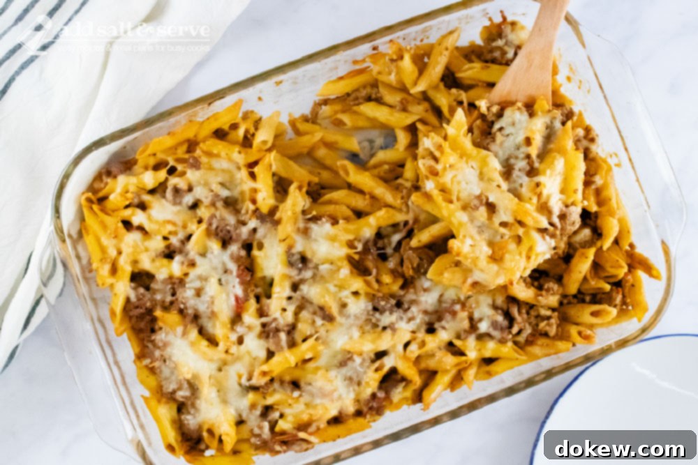 Hearty Prep-Ahead Penne Bake 2 Overhead view of penne pasta bake topped with melted cheese in a glass pan with a spoonful scooped out and held toward the camera.