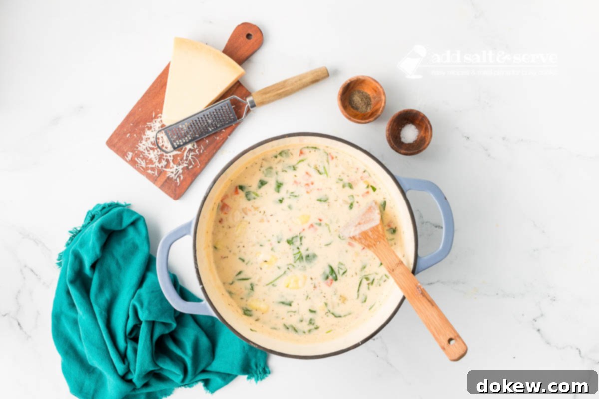 Homemade Olive Garden Chicken Gnocchi Soup 8 A large enamel Dutch oven filled with a rich, creamy Copycat Olive Garden Chicken Gnocchi Soup, with a wooden spoon resting in it. A wedge of Parmesan cheese, a salt shaker, and a pepper mill are visible beside the pot.