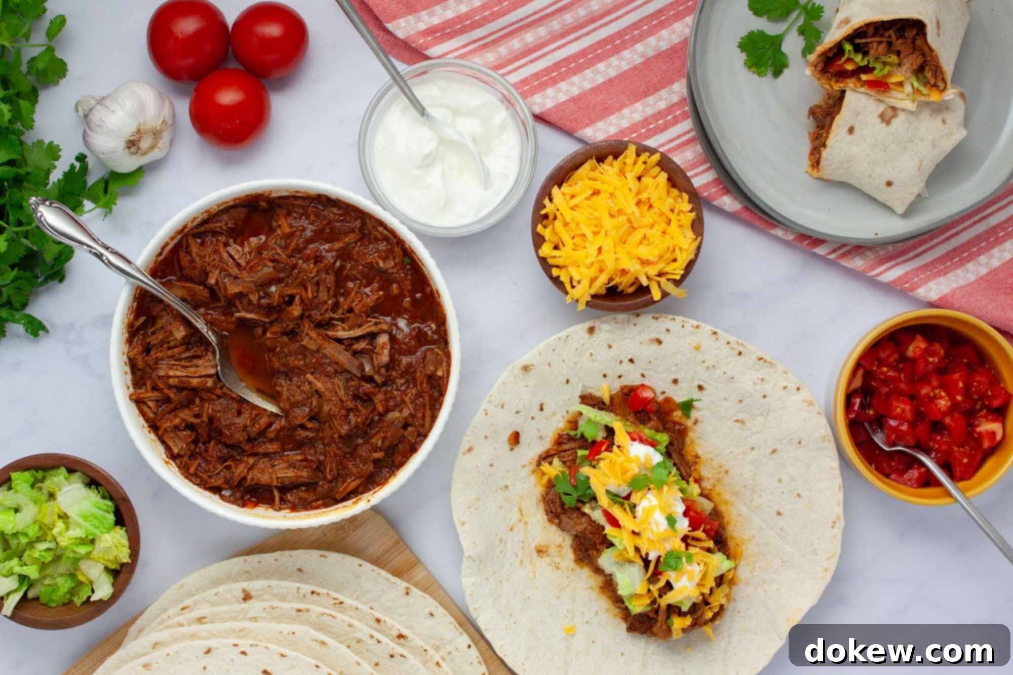 Tender Slow Cooker Beef Burritos 2 Overhead view of slow cooker beef burrito meat in a bowl surrounded by garnishes, tortillas, and a plate with a half-assembled burrito.