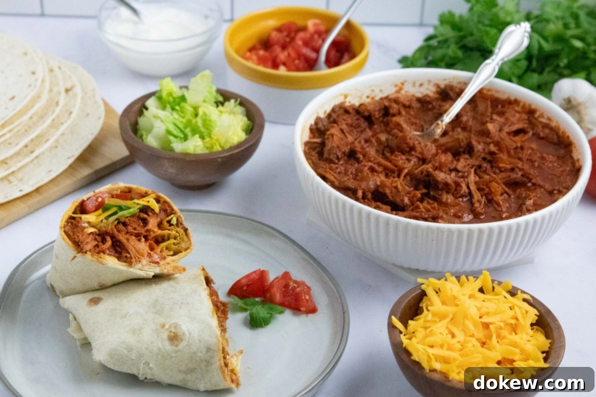Tender Slow Cooker Beef Burritos 7 Overhead view of slow cooker beef burrito meat in a bowl surrounded by garnishes, tortillas, and a plate with an assembled burrito.
