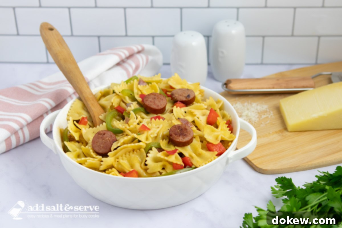 Fast & Flavorful Kielbasa Pepper Pasta 2 Bowtie pasta with red and green bell peppers, red onion, and sliced kielbasa with a spoon in a serving dish beside fresh parsley and Parmesan cheese.