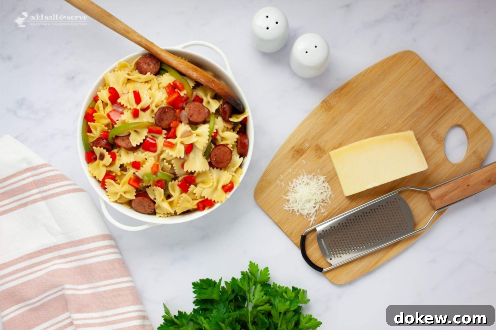 Fast & Flavorful Kielbasa Pepper Pasta 6 Bowtie pasta with red and green bell peppers, red onion, and sliced kielbasa with a spoon in a serving dish beside fresh parsley and Parmesan cheese.
