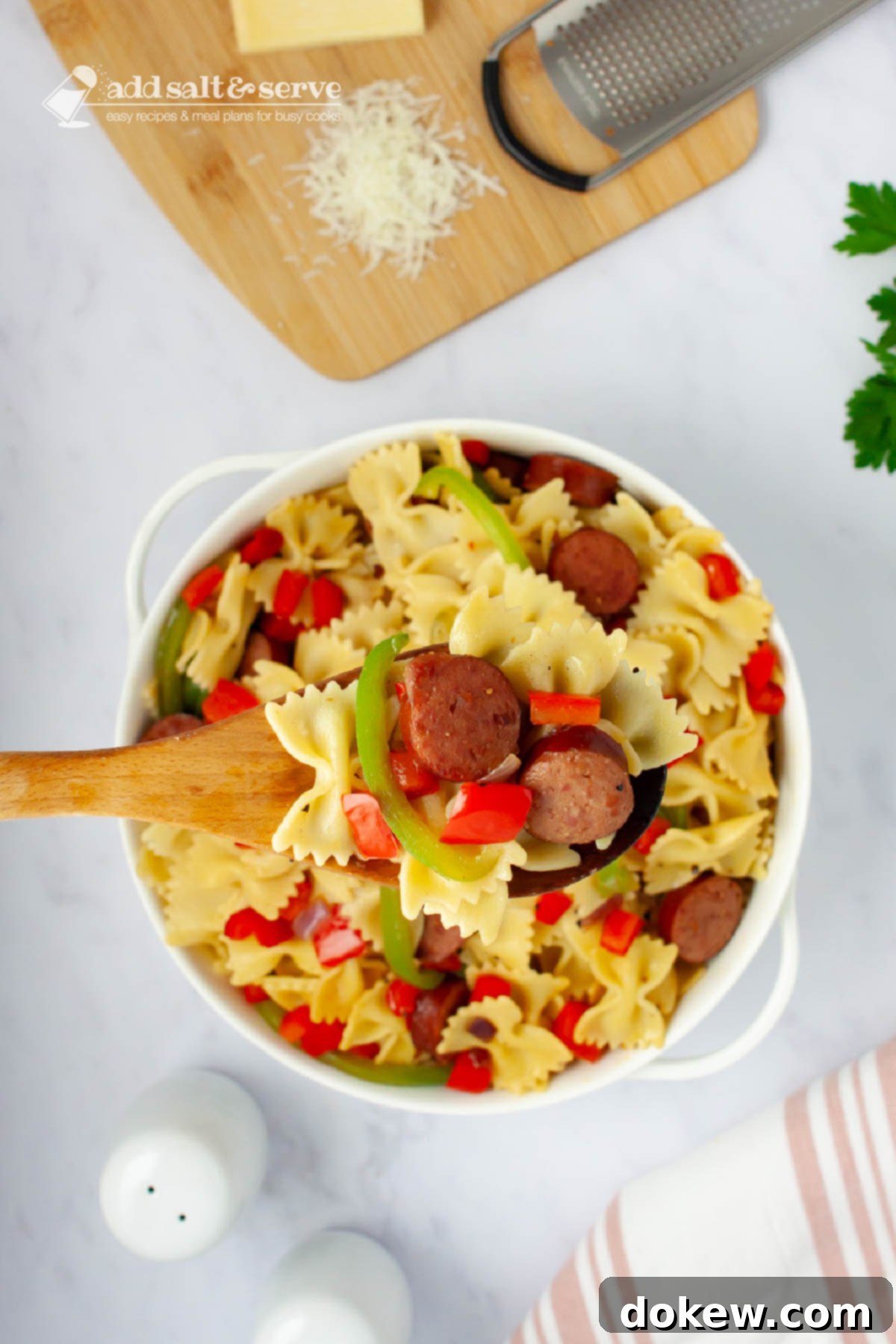 Fast & Flavorful Kielbasa Pepper Pasta 7 Bowtie pasta with red and green bell peppers, red onion, and sliced kielbasa with a spoon in a serving dish beside fresh parsley and Parmesan cheese.