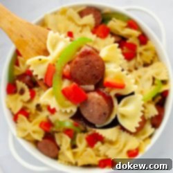Fast & Flavorful Kielbasa Pepper Pasta 8 A wooden spoonful of bowtie pasta with red and green bell peppers, red onion, and sliced kielbasa held above a service dish of the same.
