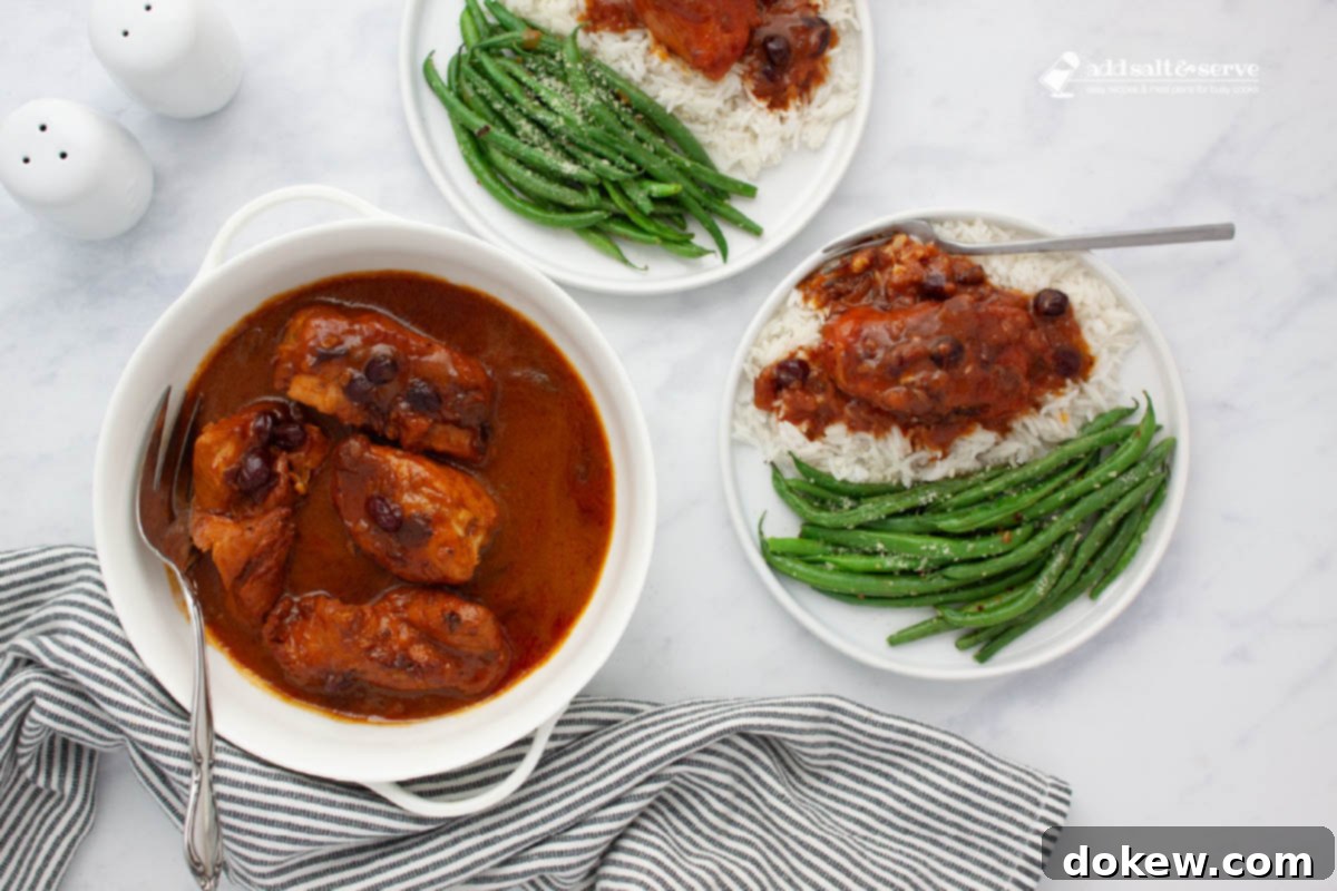 Slow Cooker Cranberry French Glazed Chicken 2 White serving dish presenting cooked cranberry chicken breasts in a vibrant sauce, with whole cranberries visible. The dish is accompanied by a serving fork, and two plates feature seasoned green beans and chicken served over rice, all arranged on a kitchen counter.