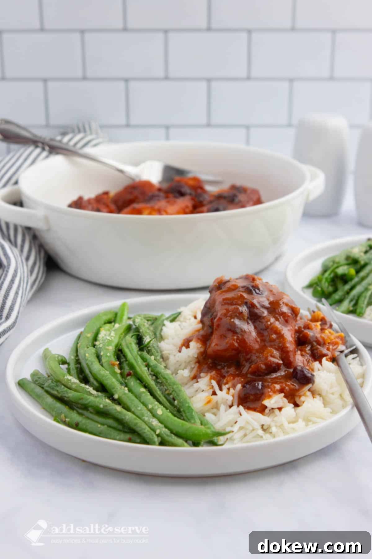 Slow Cooker Cranberry French Glazed Chicken 6 A beautifully plated dish featuring tender chicken in a reddish-orange cranberry sauce, served over white rice with a side of seasoned green beans. In the background, a serving dish of the same chicken and sauce is visible, highlighting a delicious and satisfying meal.