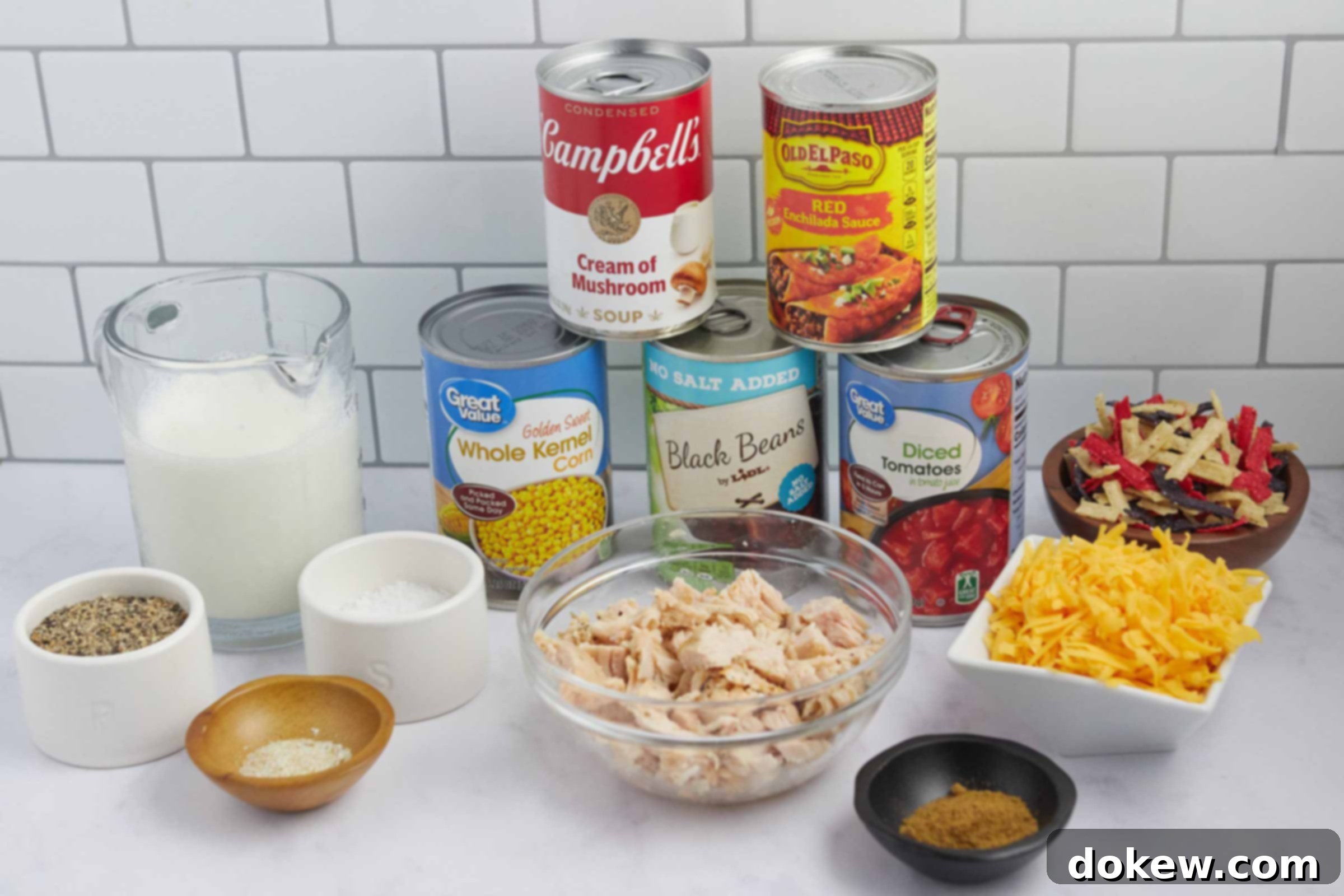 Velvety 15-Minute Chicken Enchilada Soup 3 A flat lay photograph showcasing all the ingredients required for Creamy Chicken Enchilada Soup: a carton of milk, a can of corn, a can of black beans, a can of cream of mushroom soup, a can of red enchilada sauce, a can of diced tomatoes, chopped rotisserie chicken, small bowls of salt, pepper, cumin, garlic salt, a pile of shredded cheddar cheese, and a handful of tortilla strips.