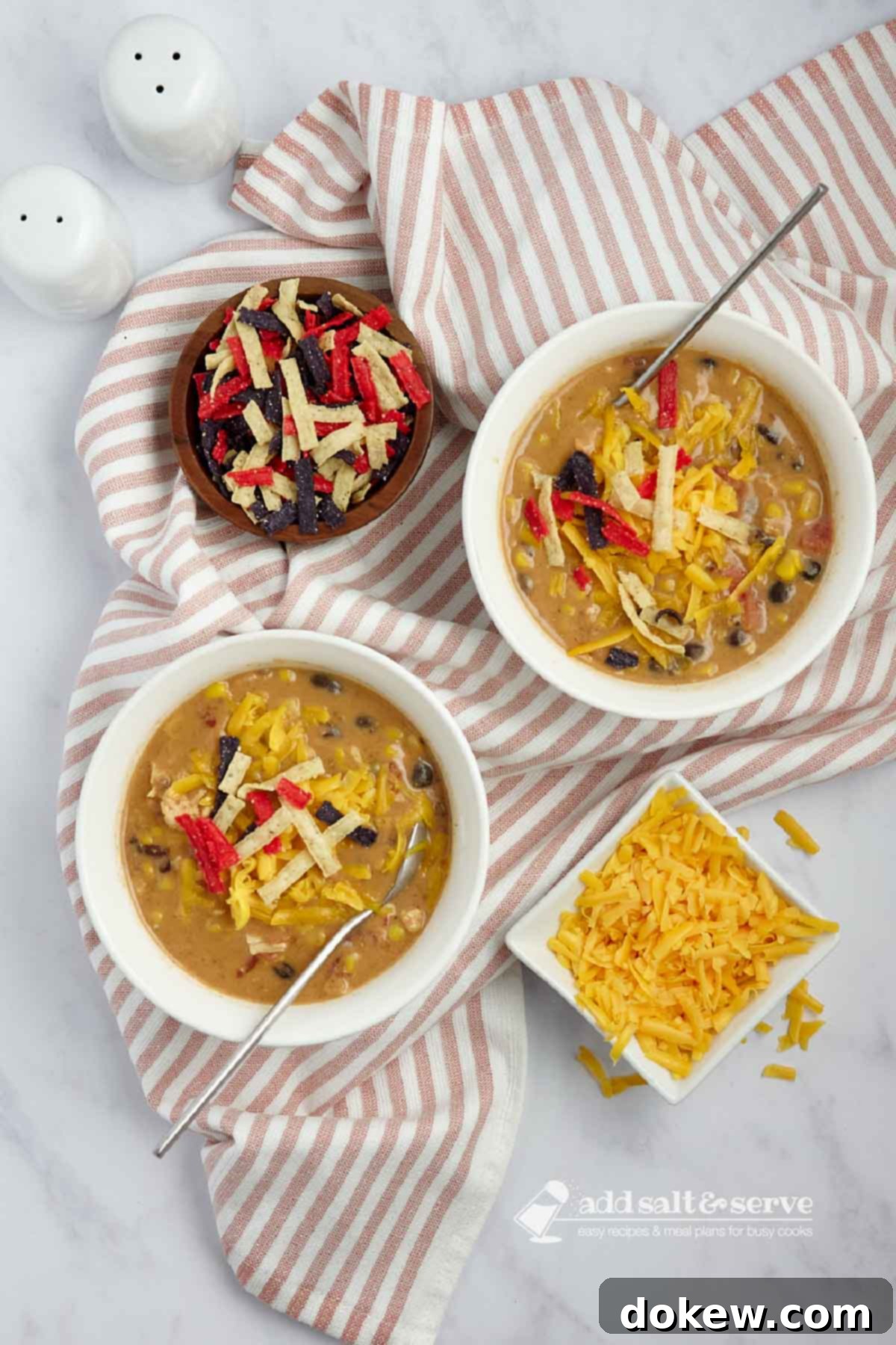 Velvety 15-Minute Chicken Enchilada Soup 6 Two bowls of chicken enchilada soup garnished with shredded cheddar cheese, fresh cilantro, and tortilla strips. Beside them are small separate bowls holding extra shredded cheddar cheese and crispy tortilla strips, ready for more topping.