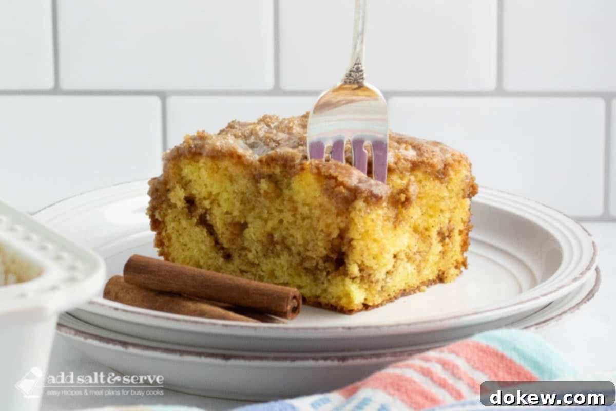 Cinnamon Honey Swirl Cake 2 A delectable slice of Honey Bun Cake on a white plate, showcasing its golden-brown crust, a rich cinnamon sugar swirl in the middle, and a glistening vanilla glaze on top. A fork is gently placed in the cake, ready for a bite.