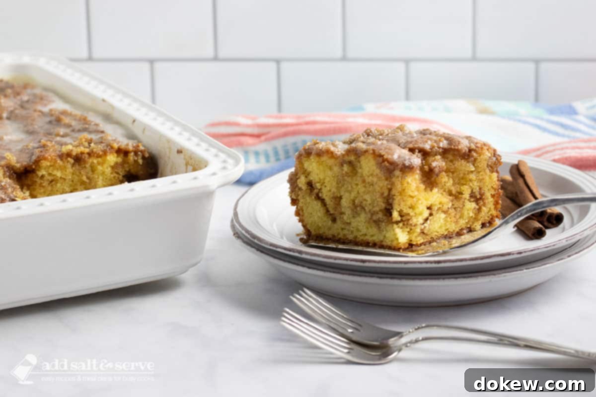 Cinnamon Honey Swirl Cake 7 A perfectly baked Honey Bun Cake sits in its pan on a kitchen counter, with a slice being removed and placed onto a plate. The rich cinnamon swirl and smooth vanilla glaze are clearly visible, with two forks resting nearby, inviting enjoyment.