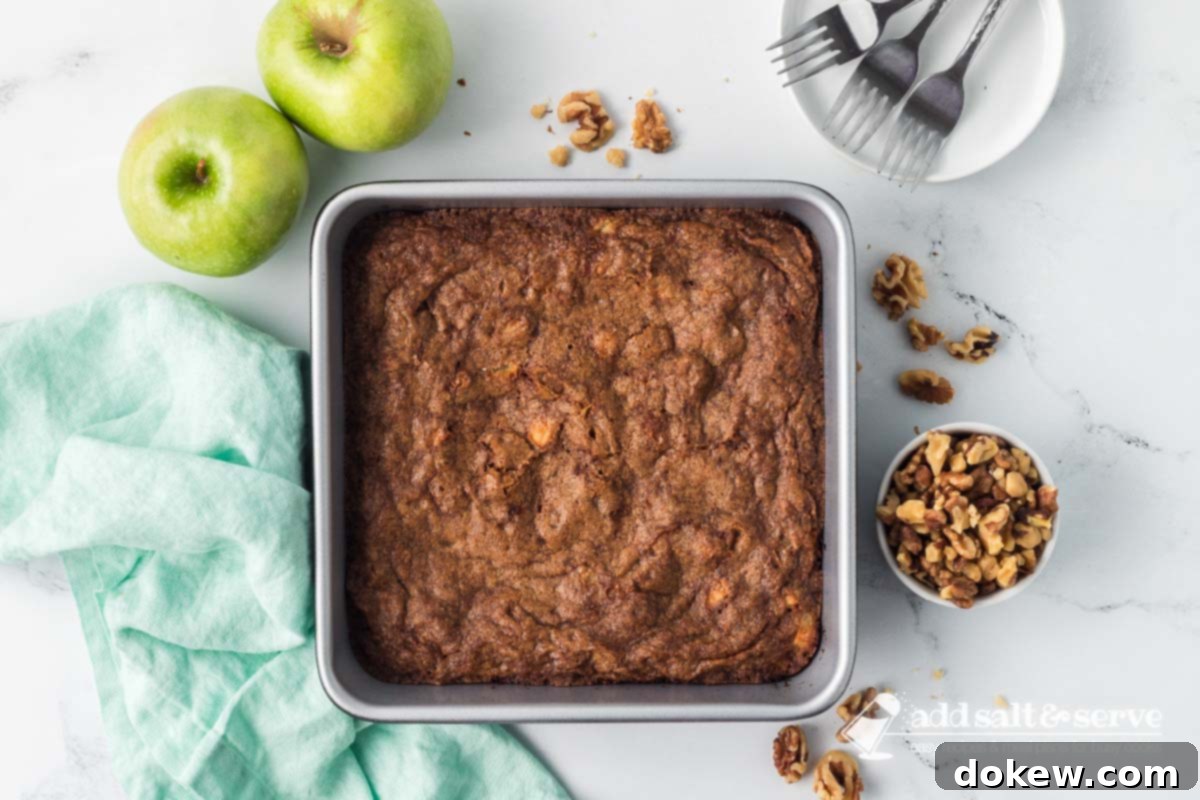 Spiced Apple Walnut Bars 9 Baked Apple Walnut Bars in an 9-inch by 9-inch pan on a counter surrounded by two green apples, a bowl of walnuts, and a white plate with three forks.