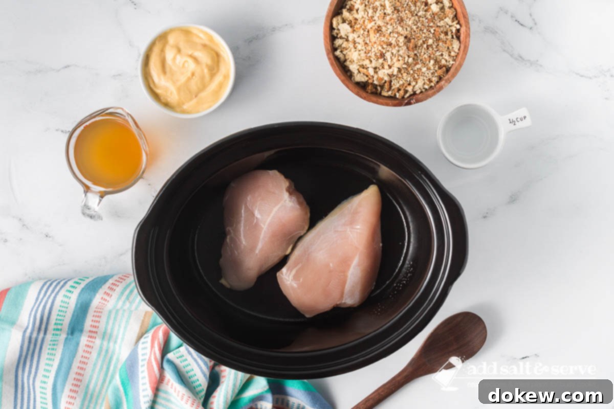 Effortless Slow Cooker Chicken and Stuffing 4 Initial setup in a crockpot: two uncooked boneless chicken breasts, surrounded by bowls of dressing mix, cream of chicken soup, chicken broth, and water.