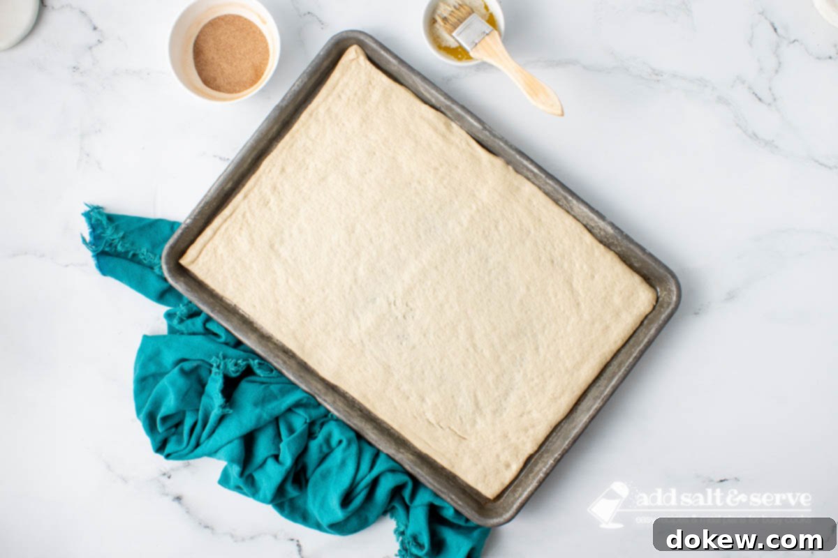 Quick Pizza Dough Cinnamon Twists 5 Unrolled pizza crust dough laid flat on a greased baking sheet with a bowl of melted butter and the cinnamon sugar mixture nearby.