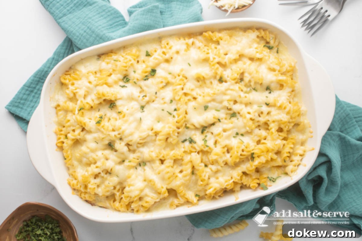 Golden Baked Alfredo Rotini 12 Rotini Pasta Bake in a baking dish resting on a tea towel with forks, shredded cheese, and chopped fresh parsley surrounding the pan.