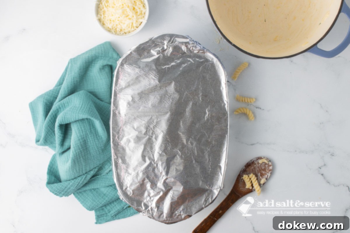 Golden Baked Alfredo Rotini 10 An oblong baking dish covered in aluminum foil on a counter beside an empty Dutch oven and a wooden spoon with saucy rotini on it.