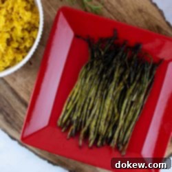 Perfectly Grilled Asparagus 2 grilled asparagus on a red serving plate placed on a wood serving tray with a bowl of yellow rice and a tea towel to the side