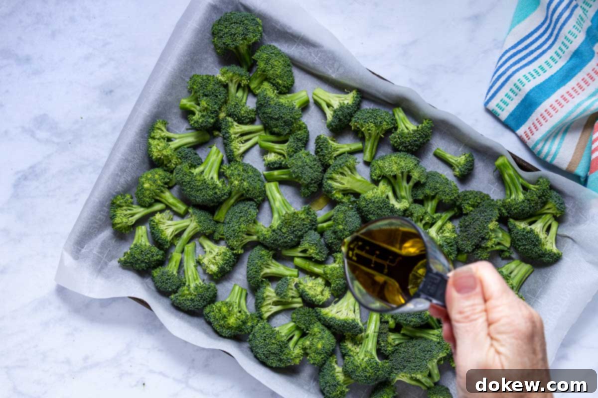 Crispy Garlic Roasted Broccoli 7 A person gently drizzles extra virgin olive oil over a single layer of fresh broccoli florets on a parchment-lined baking sheet using a small measuring cup.