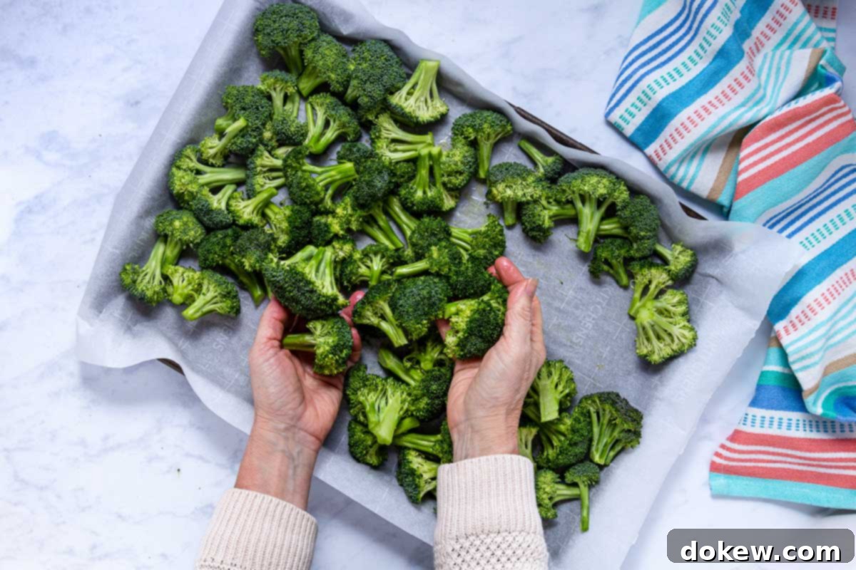 Crispy Garlic Roasted Broccoli 8 A person uses their hands to gently toss and coat broccoli florets with olive oil and seasonings directly on a parchment-lined baking sheet, ensuring even distribution.