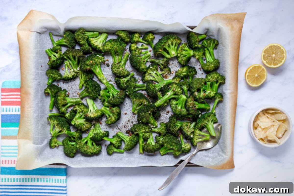Crispy Garlic Roasted Broccoli 9 A baking sheet filled with perfectly roasted broccoli florets, some slightly charred, is presented alongside a small bowl of shaved Parmesan cheese and two lemon halves, ready for the final touches.