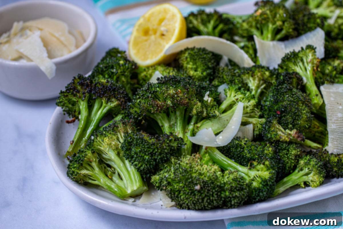 Crispy Garlic Roasted Broccoli 10 A serving of roasted broccoli florets, garnished with a vibrant lemon half and a sprinkle of Parmesan cheese, presented on a white plate, ready to be enjoyed.