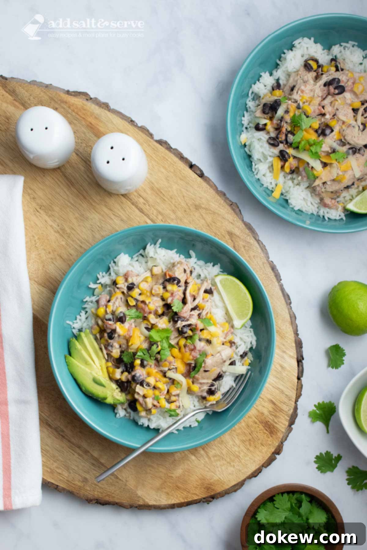 Easy Slow Cooker Fiesta Chicken 6 Bowl of shredded chicken in a creamy sauce with black beans, corn, tomatoes, and green chilies over rice and garnished with sliced avocado, shredded cheese, fresh cilantro, and lime wedges.