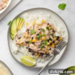 Easy Slow Cooker Fiesta Chicken 7 A plate of shredded chicken in a creamy sauce with black beans, corn, tomatoes, and green chilies served over rice and garnished with sliced avocado, shredded cheese, fresh cilantro, and lime wedges.