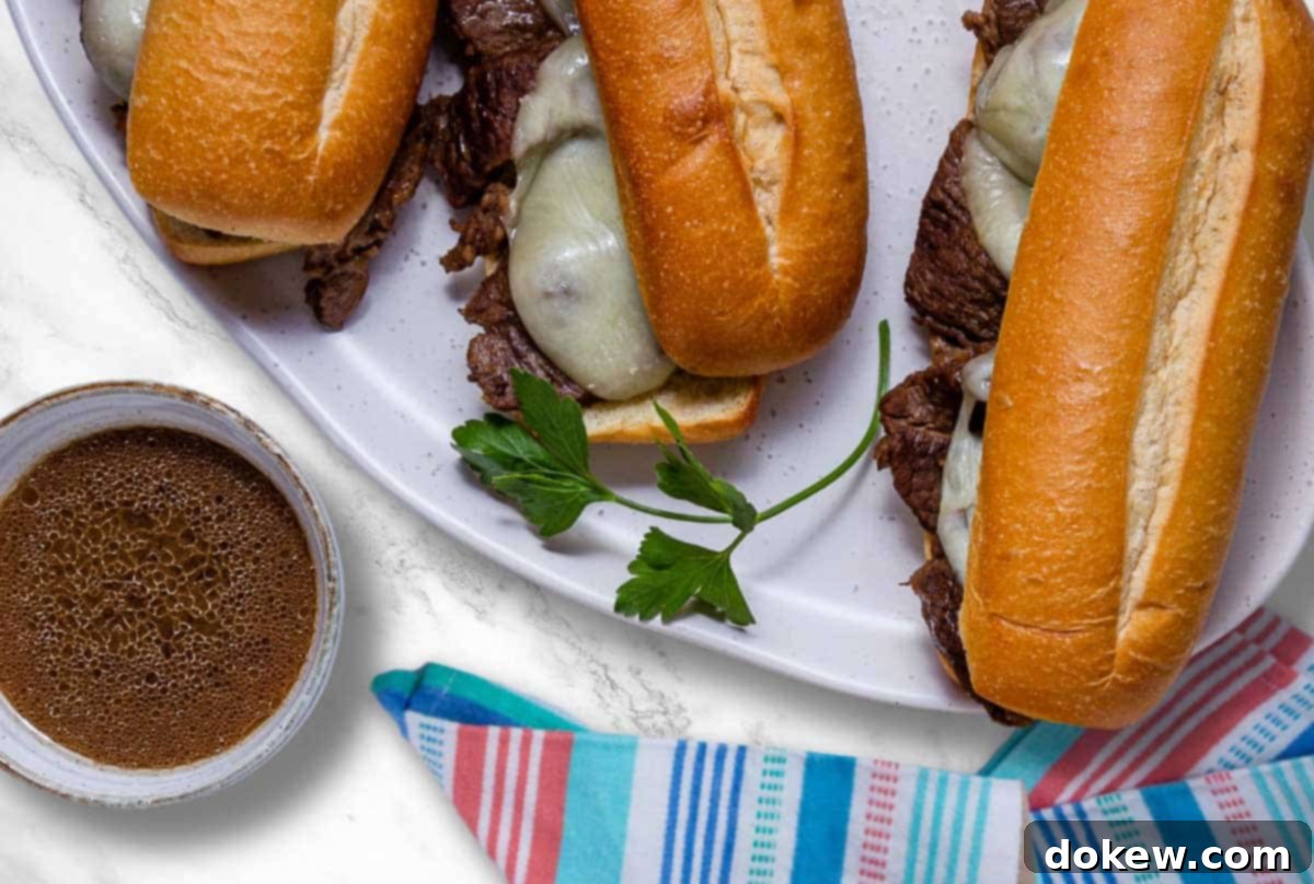 Easy Crockpot French Dip Tender Beef and Flavorful Au Jus 2 A plate of French Dip sandwiches beside a bowl of au jus, showcasing the tender beef and melted cheese on a toasted hoagie roll.