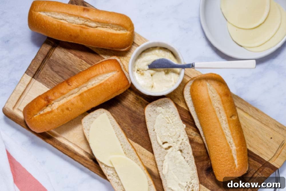 Easy Crockpot French Dip Tender Beef and Flavorful Au Jus 5 Hoagie rolls sliced lengthwise on a cutting board, with one side spread with horseradish sauce and the other topped with provolone cheese.