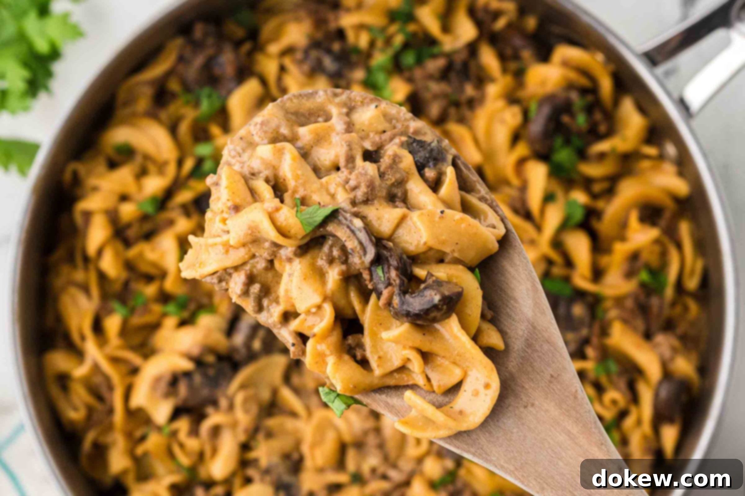 A pan filled with creamy Ground Beef Stroganoff, featuring tender egg noodles, savory mushrooms, and ground beef, garnished with fresh chopped parsley. A wooden spoon lifts a serving toward the camera.
