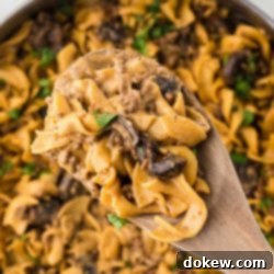 Simple One-Pan Creamy Ground Beef Stroganoff 11 A pan with a finished recipe showing egg noodles, sliced mushrooms, and ground beef in a sauce garnished with chopped parsley with a serving lifted toward the camera on a wooden spoon.
