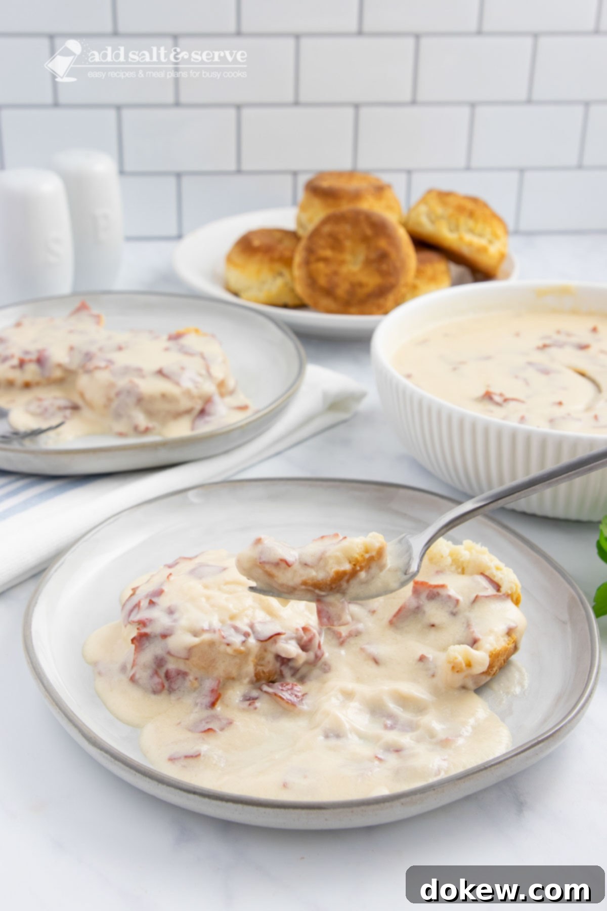 Hearty Chipped Beef Gravy 7 Plate of creamy chipped beef gravy over two biscuit halves with a forkful held above the plate on a counter next to another plate of biscuits and gravy, a bowl of the gravy, and a plate of biscuits, showing a delightful breakfast spread.