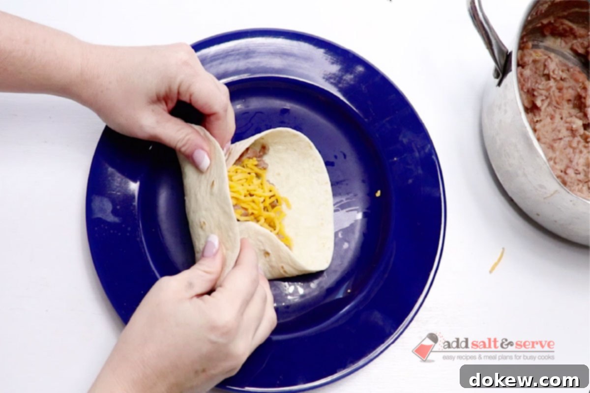 A tortilla with bean burrito filling and shredded cheddar cheese being folded into a burrito.