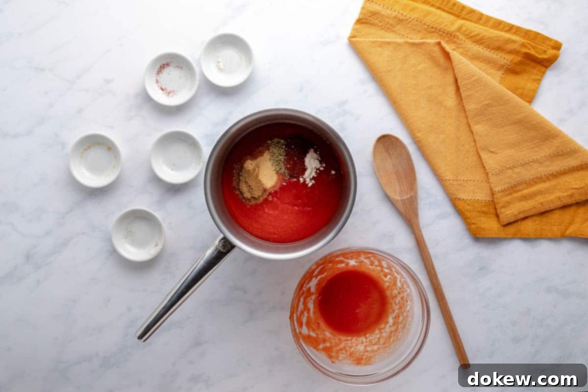 Effortless Homemade Enchilada Sauce 3 Tomato sauce, chili powder, ground cumin, onion powder, garlic powder, and dried oregano in a saucepan with a wooden spoon and a tea towel beside it on the counter.