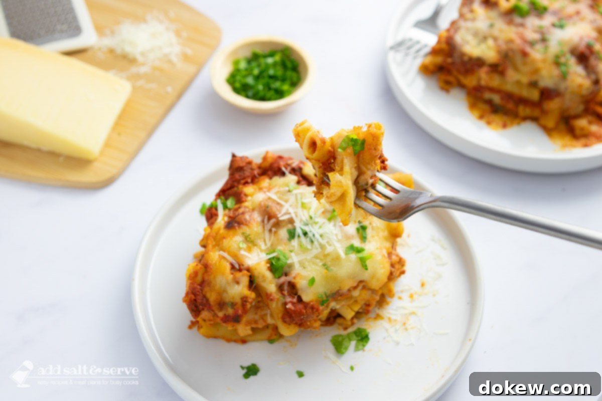 Classic Cheesy Meatless Baked Ziti 2 Two generous servings of cheesy meatless baked ziti in white bowls, accompanied by a wedge of Parmesan cheese on a wooden cutting board. The main baking pan with more ziti is visible in the soft-focus background.