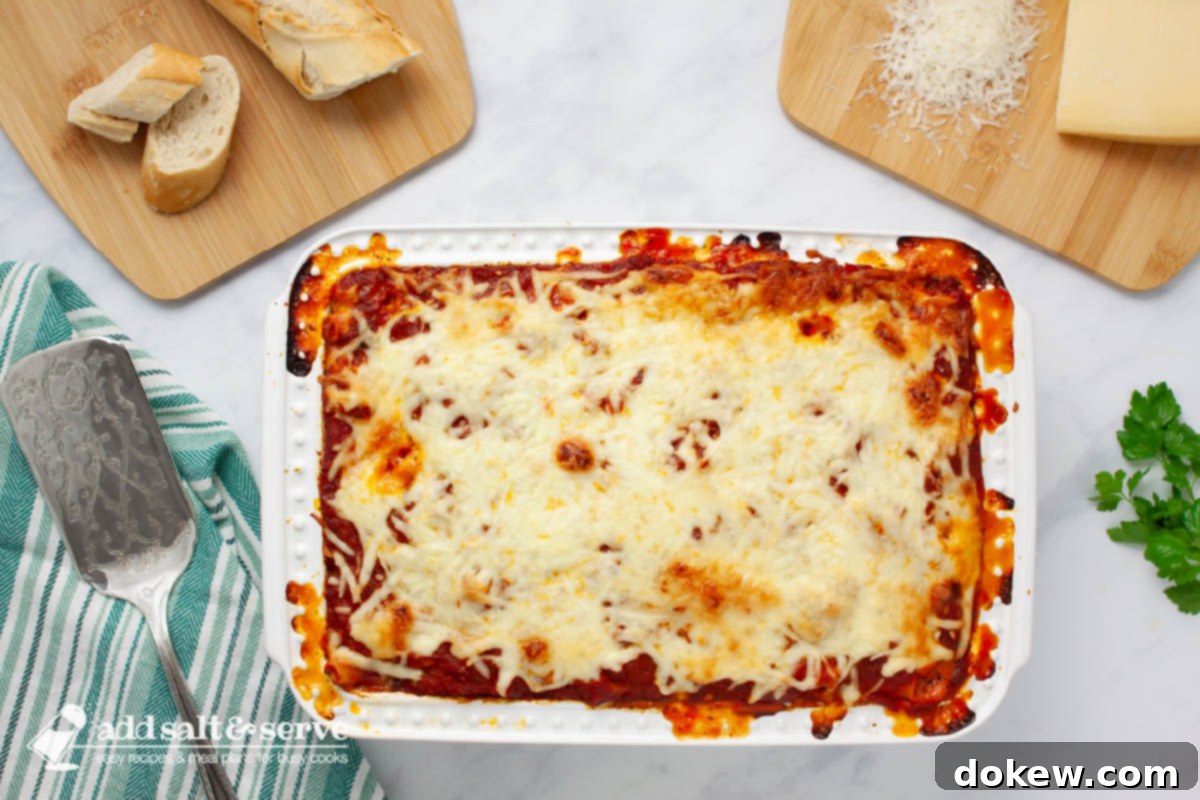 Classic Cheesy Meatless Baked Ziti 8 A freshly baked pan of cheesy meatless baked ziti, garnished with sprigs of fresh parsley, placed on a kitchen counter. Slices of baguette bread and a block of Parmesan cheese are visible nearby, creating an inviting scene.