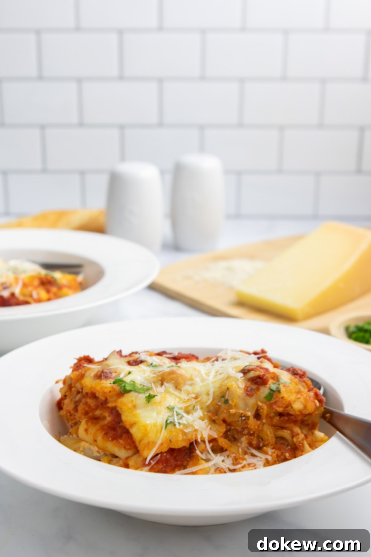 Classic Cheesy Meatless Baked Ziti 9 A single serving of cheesy meatless baked ziti in a white bowl, with a fork poised to take a bite. A wooden cutting board with Parmesan cheese is in the background.