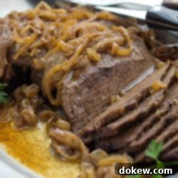 Tender Slow Cooker London Broil in Savory Au Jus 9 Slices of beef on a white platter. Beef is covered with cooked onions and resting in an au jus. The plate is garnished with parsley and there is a steak knife resting on the back of the platter.