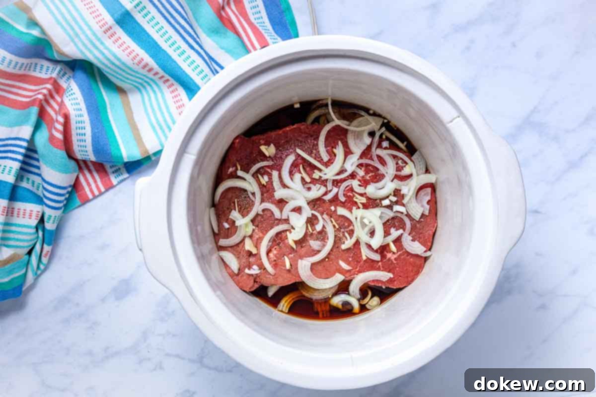 Tender Slow Cooker London Broil in Savory Au Jus 7 A crockpot with layers of sliced onions and seasoned uncooked London broil roast covered in soy sauce and water.