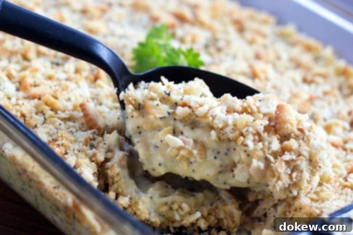 A black spoon is scooping golden-brown Poppy Seed Chicken Casserole from a clear glass casserole dish, showcasing its creamy interior and crunchy topping.