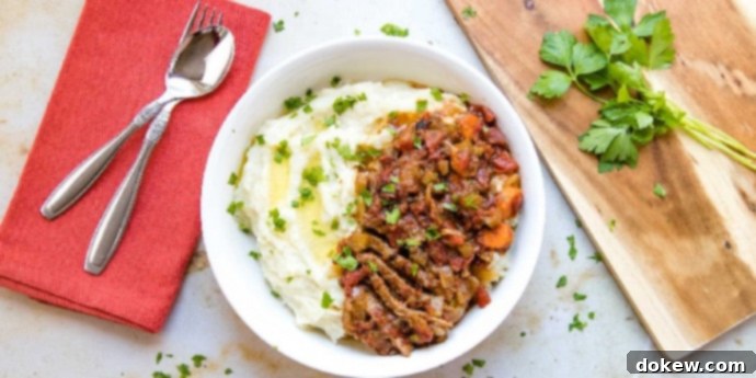 Savory Swiss Steak White bowl with mashed potatoes in one half and strips of beef and chopped vegetables in the other half, garnished with parsley. To the left of the bowl is a spoon and fork on a red linen napkin. To the right is a wooden cutting board with parsley on it.
