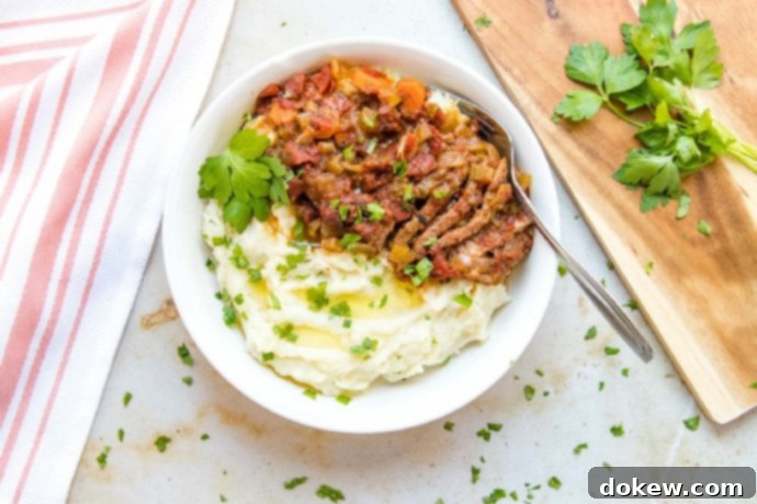 Savory Swiss Steak White bowl with mashed potatoes in one half and strips of beef and chopped vegetables in the other half, garnished with parsley. To the left of the bowl is a spoon and fork on a red linen napkin. To the right is a wooden cutting board with parsley on it.